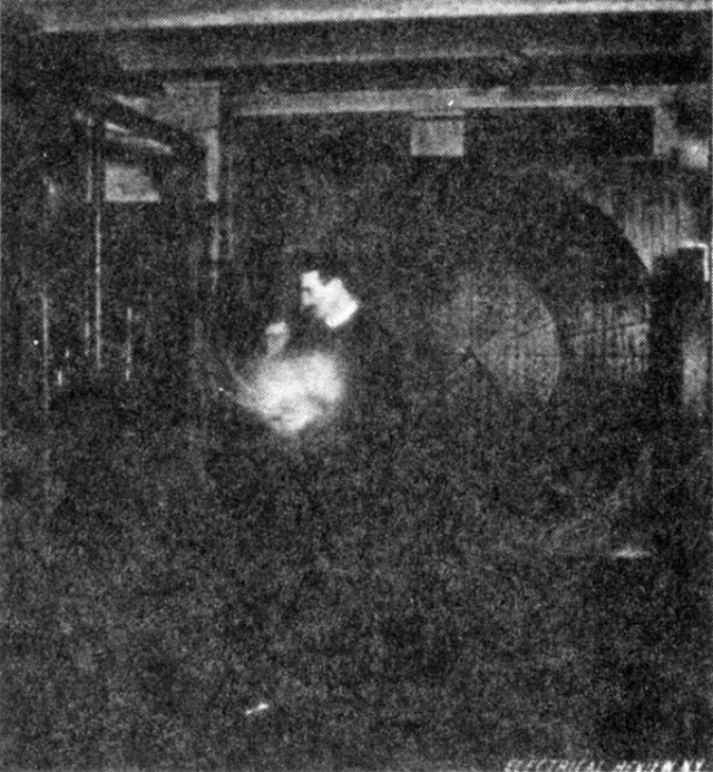 Tesla stands in the middle of the laboratory lighting a vacuum bulb with waves from a distant oscillator. In this case, his body is exposed to excessive electrical pressure.